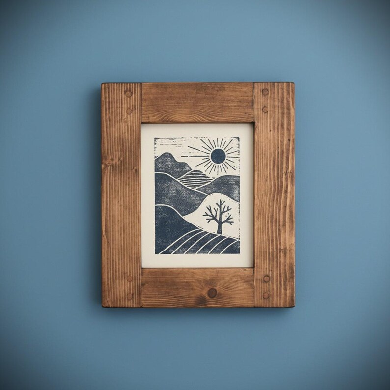 May include: A framed print depicting a landscape with hills, a tree, and a sun. The artwork uses blue and white tones, encased in a rectangular wooden frame. The frame is made of natural wood and is mounted on a blue wall.