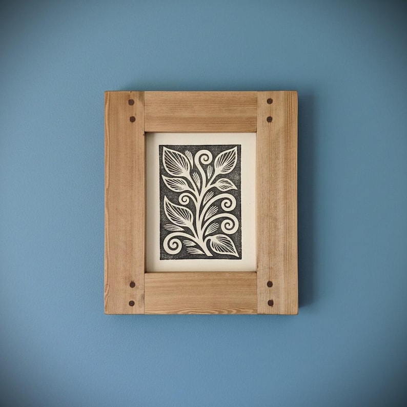 May include: A framed botanical print featuring a black and white floral design. The artwork is set within a natural wood frame, secured with small, dark-colored pegs. The frame is rectangular and mounted on a blue wall.