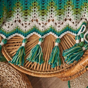 May include: A handmade crochet blanket in shades of teal, green, and cream. The blanket features a chevron pattern and is adorned with tassels in matching colors. The blanket is draped over a wicker chair.