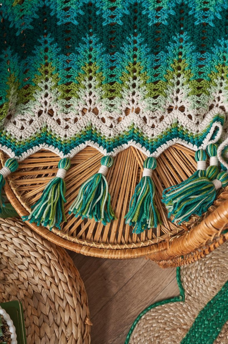 May include: A handmade crochet blanket in shades of teal, green, and cream. The blanket features a chevron pattern and is adorned with tassels in matching colors. The blanket is draped over a wicker chair.