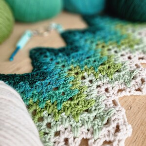 May include: A close-up of a crochet project in progress, featuring a gradient of green and blue yarn. The project is a textured, open-weave design. Several balls of yarn in various shades of green and blue are in the background, along with a crochet hook.