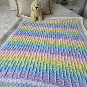 Puede incluir: Una manta rectangular de ganchillo con rayas horizontales en tonos pastel de amarillo, rosa, azul y verde. La manta presenta un patrón de punto acanalado texturizado y está rematada con un borde blanco sólido en los cuatro lados.