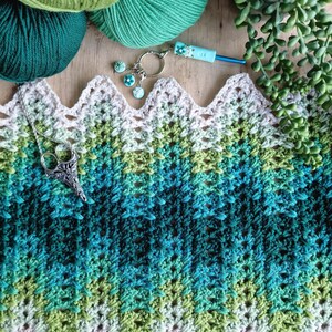 May include: A crochet project in shades of green and blue, with yarn balls, a crochet hook, and a decorative keychain. The project features a scalloped edge and a gradient color scheme. A silver pendant with scissors is also present.