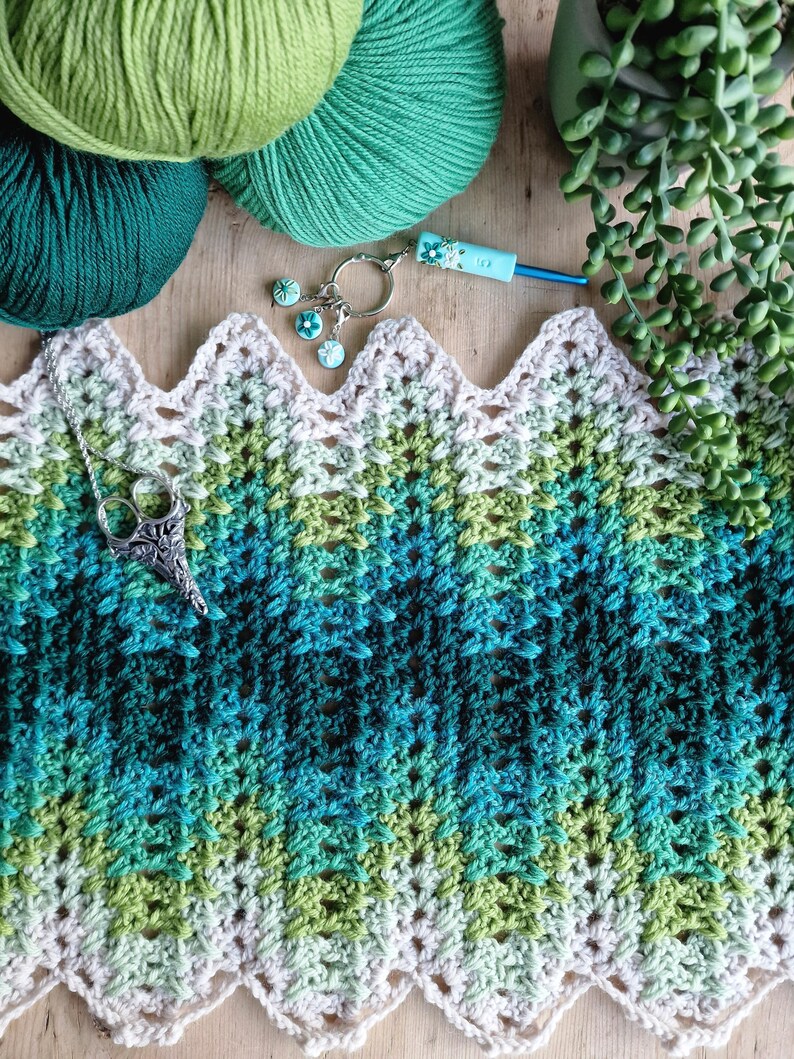 May include: A crochet project in shades of green and blue, with yarn balls, a crochet hook, and a decorative keychain. The project features a scalloped edge and a gradient color scheme. A silver pendant with scissors is also present.