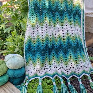 May include: A handmade crochet scarf with a chevron pattern in shades of white, green, and teal. The scarf has a scalloped edge and is adorned with matching tassels. Several balls of yarn in coordinating colors are visible.