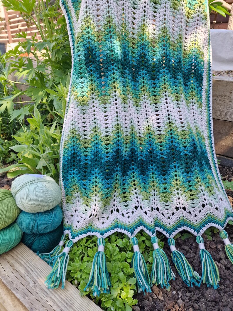May include: A handmade crochet scarf with a chevron pattern in shades of white, green, and teal. The scarf has a scalloped edge and is adorned with matching tassels. Several balls of yarn in coordinating colors are visible.