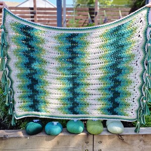 May include: A handmade crochet blanket with a chevron pattern in shades of green, teal, and white. The blanket has a scalloped edge and is adorned with green tassels. Five balls of yarn in coordinating colors are displayed below the blanket.