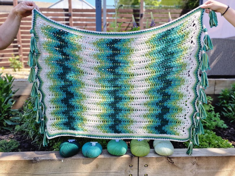 May include: A handmade crochet blanket with a chevron pattern in shades of green, teal, and white. The blanket has a scalloped edge and is adorned with green tassels. Five balls of yarn in coordinating colors are displayed below the blanket.