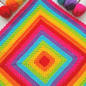 May include: A square crochet blanket featuring concentric diamond patterns in vibrant rainbow colors including red, orange, yellow, lime green, turquoise, purple, and hot pink. Several matching yarn skeins and a crochet hook are positioned nearby.