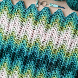 May include: A handmade crochet blanket in shades of white, green, and teal, with a zigzag edge. The blanket is surrounded by balls of yarn in matching colors, a crochet hook, and a small charm.