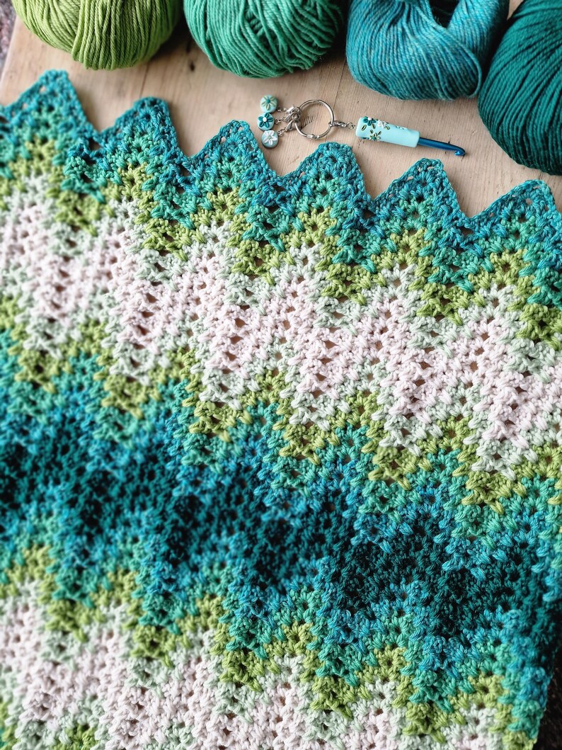 May include: A handmade crochet blanket in shades of white, green, and teal, with a zigzag edge. The blanket is surrounded by balls of yarn in matching colors, a crochet hook, and a small charm.