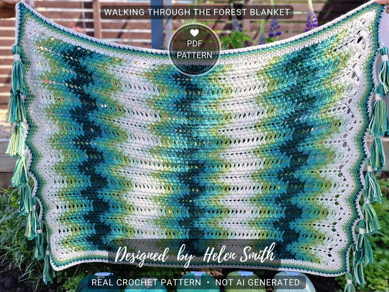 May include: A handmade crochet blanket with a chevron pattern in shades of white, teal, and green. The blanket has a scalloped edge and is adorned with tassels at each corner. Text on the blanket reads "WALKING THROUGH THE FOREST BLANKET" and "Designed by Helen Smith".
