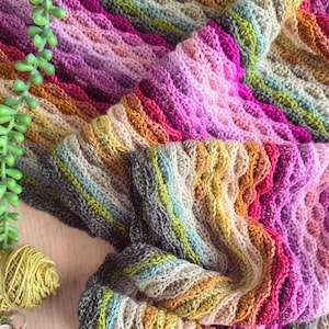 Peut inclure: Une couverture crochetée présentant un motif de vagues en relief avec un dégradé de couleurs vives, notamment des tons de rose, magenta, violet, jaune, orange, vert et gris. Le textile présente une surface texturée et ondulée sur l'ensemble.