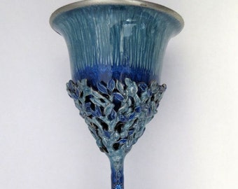 May include: A blue ceramic goblet with a decorative, leafy design around the bowl and a textured, blue stem. The base of the goblet is wide and flared.