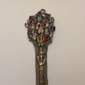 May include: A gold-toned metal mezuzah with a tree of life design. The tree has red and green leaves and is decorated with gold accents. The mezuzah is a traditional Jewish religious item that is attached to the doorpost of a home.