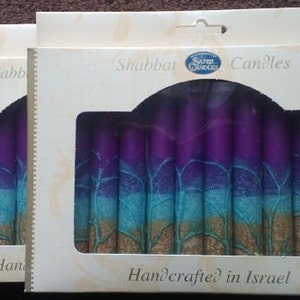 Puede incluir: Dos cajas de velas de Shabat con un dise&ntilde;o morado, azul y dorado. Las cajas est&aacute;n etiquetadas como "Shabbat Candles" y "Handcrafted in Israel".
