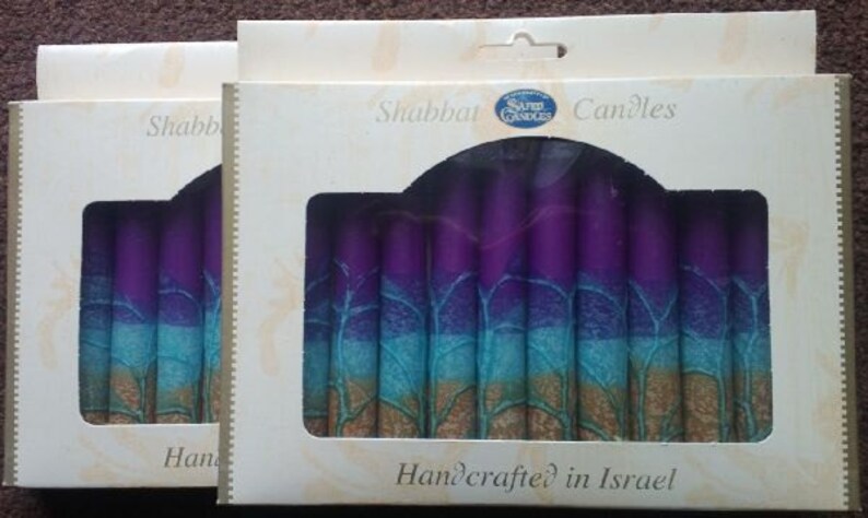Puede incluir: Dos cajas de velas de Shabat con un dise&ntilde;o morado, azul y dorado. Las cajas est&aacute;n etiquetadas como "Shabbat Candles" y "Handcrafted in Israel".