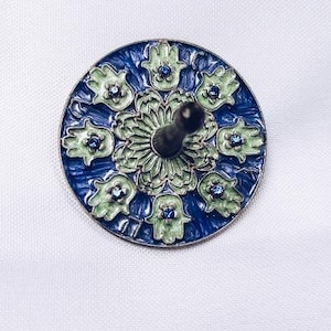 May include: A blue and green metal dreidel with a hamsa hand design. The dreidel has a black spinning top.