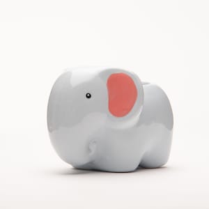 Puede incluir: Un macetero de cerámica gris claro en forma de elefante con una oreja rosa. El macetero tiene un pequeño ojo negro y una forma redondeada.