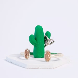 May include: A green cactus-shaped ring holder with two gold rings and a silver ring on it. The holder is sitting on a white hexagonal base.