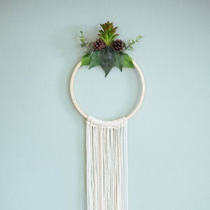 May include: A white macrame wall hanging with a wooden hoop and a green plant with brown pinecones at the top.