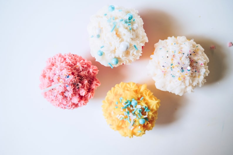 May include: Four frosted cupcakes with colorful sprinkles. The cupcakes are pink, white, yellow, and white.