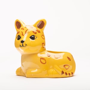 Könnte beinhalten: Ein gelber Keramik-Pflanztopf in Form eines Comic-Leoparden mit braunen Flecken. Der Topf hat eine breite, offene Oberseite und einen kleinen, abgerundeten Boden.