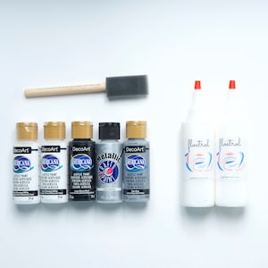 Puede incluir: Un conjunto de suministros para manualidades para pintar, que incluye cinco botellas de pintura acrílica en blanco, negro y plateado, una botella de pintura artesanal metálica, dos botellas de floetrol y un pincel de espuma.