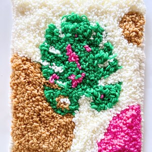 Puede incluir: Un tapiz de pared cuadrado y texturizado con un diseño afelpado. La obra de arte presenta una forma central verde y frondosa con acentos rosas, rodeada de secciones crema, marrón claro y fucsia. La pieza tiene una estética artesanal y acogedora.