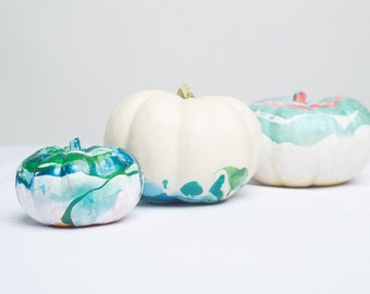 May include: Three white pumpkins painted with blue, green, and white marbled paint. The pumpkins are arranged in a row from smallest to largest.