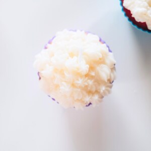 May include: A white cupcake with purple frosting in a purple cupcake liner. The cupcake is on a white surface.