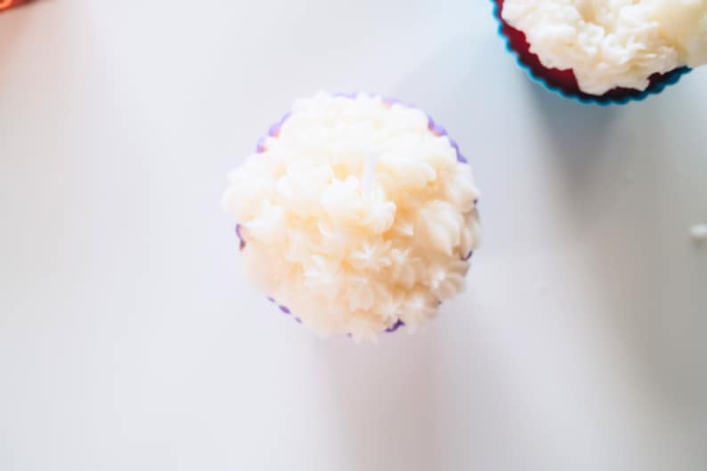 May include: A white cupcake with purple frosting in a purple cupcake liner. The cupcake is on a white surface.