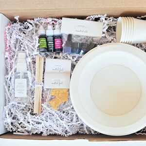 May include: A box filled with supplies for making candles, including wax flakes, essential oils, wicks, and a white bowl. The box also includes a bottle of fragrance oil, a spray bottle, and a set of paper cups.