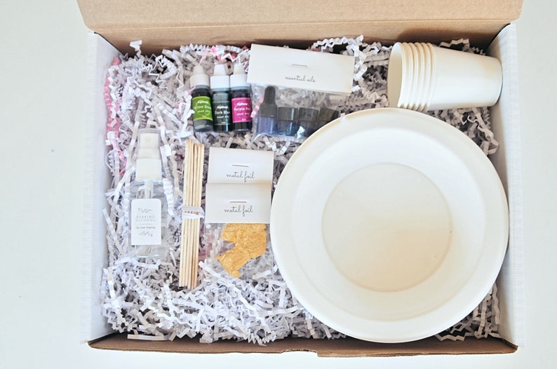 May include: A box filled with supplies for making candles, including wax flakes, essential oils, wicks, and a white bowl. The box also includes a bottle of fragrance oil, a spray bottle, and a set of paper cups.