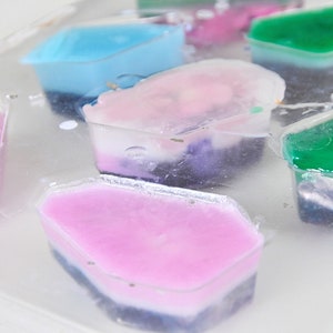 May include: A close-up of a tray of colorful, layered soap bars. The bars are in various shapes and sizes, and each has a different color combination. The soap bars are made with natural ingredients and are perfect for a relaxing bath or shower.