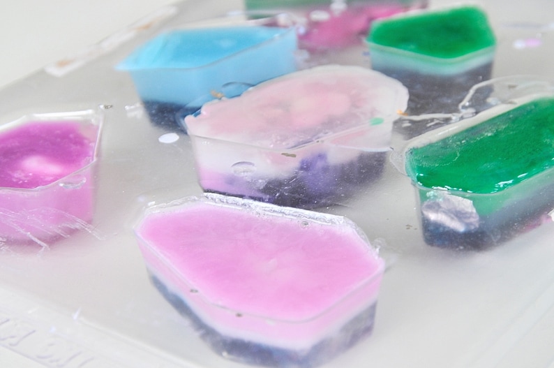 May include: A close-up of a tray of colorful, layered soap bars. The bars are in various shapes and sizes, and each has a different color combination. The soap bars are made with natural ingredients and are perfect for a relaxing bath or shower.
