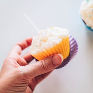 May include: A yellow cupcake liner with white frosting and colorful sprinkles. The cupcake is held in a hand and has a white candle inserted in the frosting.