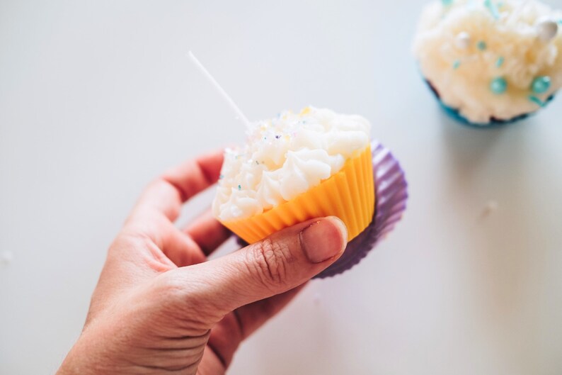 May include: A yellow cupcake liner with white frosting and colorful sprinkles. The cupcake is held in a hand and has a white candle inserted in the frosting.