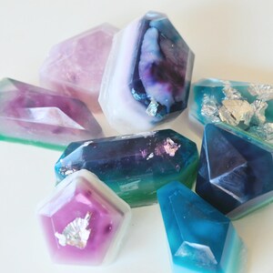 May include: A collection of eight handmade soap bars shaped like gemstones. The soaps are in shades of purple, blue, green, and white with silver flecks.