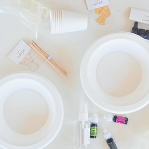 May include: A white table with two white paper plates, a bag of clear soap cubes, a bag of gold flakes, a bag of essential oils, a stack of white paper cups, a bag of clear glitter, wooden skewers, a bottle of clear spray, a bottle of green essential oil, a bottle of purple essential oil, and a bottle of black essential oil.