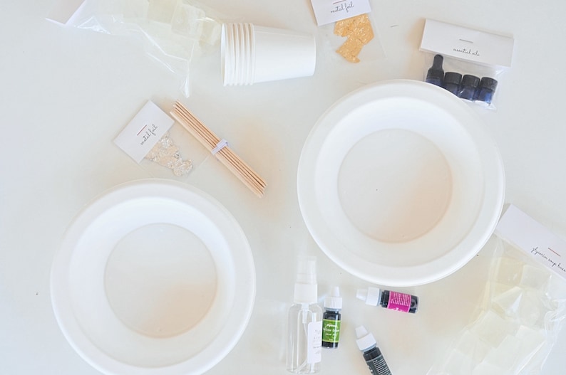 May include: A white table with two white paper plates, a bag of clear soap cubes, a bag of gold flakes, a bag of essential oils, a stack of white paper cups, a bag of clear glitter, wooden skewers, a bottle of clear spray, a bottle of green essential oil, a bottle of purple essential oil, and a bottle of black essential oil.