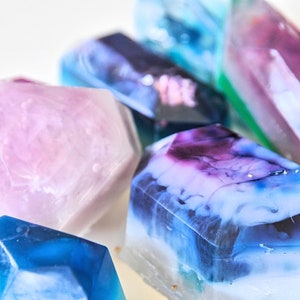 May include: A collection of colorful, geometrically shaped soap bars. The bars are translucent and have a marbled appearance. The colors include pink, blue, purple, and green.