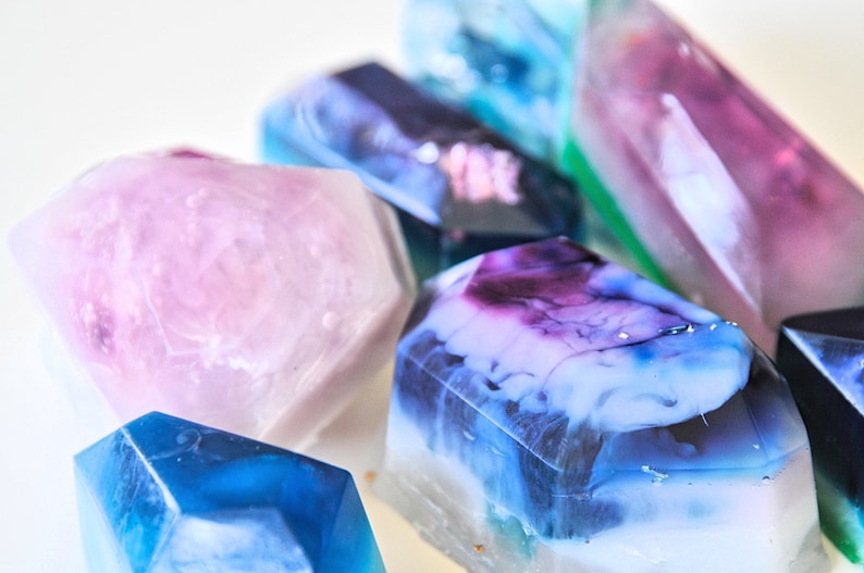 May include: A collection of colorful, geometrically shaped soap bars. The bars are translucent and have a marbled appearance. The colors include pink, blue, purple, and green.