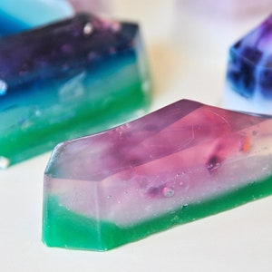 May include: A set of three layered, translucent, geometric soaps in shades of green, blue, purple, and white. The soaps are shaped like crystals and have a glossy finish.