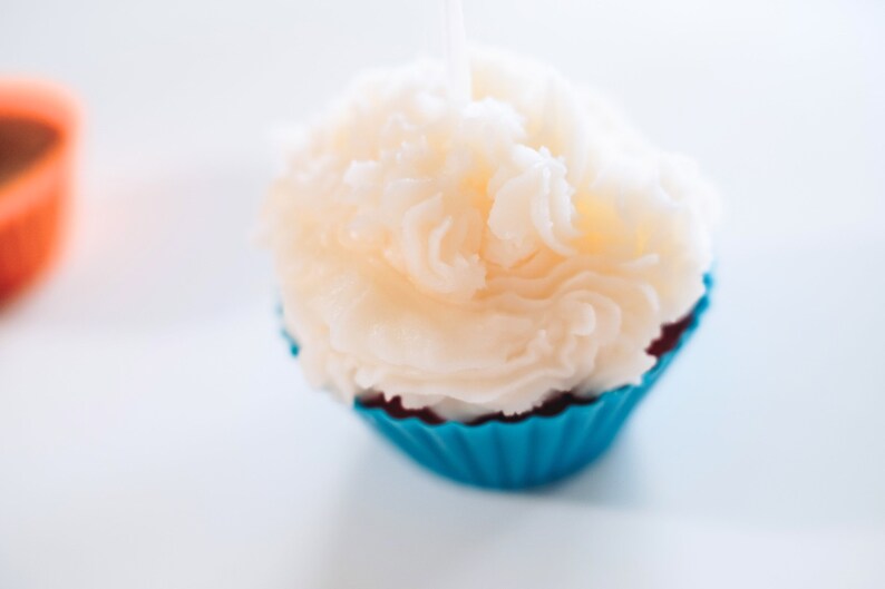 May include: A white cupcake candle with a blue and red cupcake liner. The candle is shaped like a cupcake with white frosting.