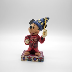 Op de afbeelding: Mickey Mouse-beeldje als tovenaar, met een blauwe hoed met gele sterren en een gele toverstaf. De rode mantel heeft gedetailleerde patronen en het beeldje staat op een decoratieve basis.