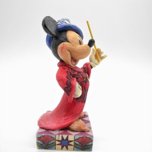 Op de afbeelding: Een Mickey Mouse beeldje in een rode mantel en blauwe hoed, met een gele toverstaf. Het beeldje staat op een decoratieve basis met een geometrisch patroon. Een verzamelobject.