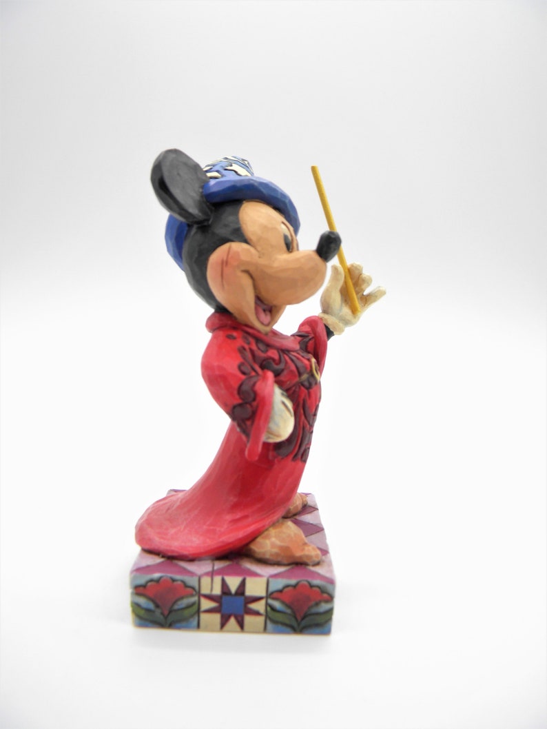 Op de afbeelding: Een Mickey Mouse beeldje in een rode mantel en blauwe hoed, met een gele toverstaf. Het beeldje staat op een decoratieve basis met een geometrisch patroon. Een verzamelobject.