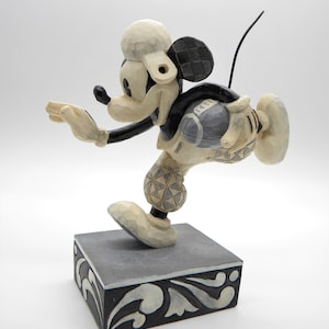 Puede incluir: Una figura tallada de Mickey Mouse en una pose din&aacute;mica, con una combinaci&oacute;n de colores crema y negro. La figura est&aacute; montada sobre una base cuadrada con adornos decorativos. El dise&ntilde;o de la figura presenta patrones geom&eacute;tricos y una est&eacute;tica caprichosa.