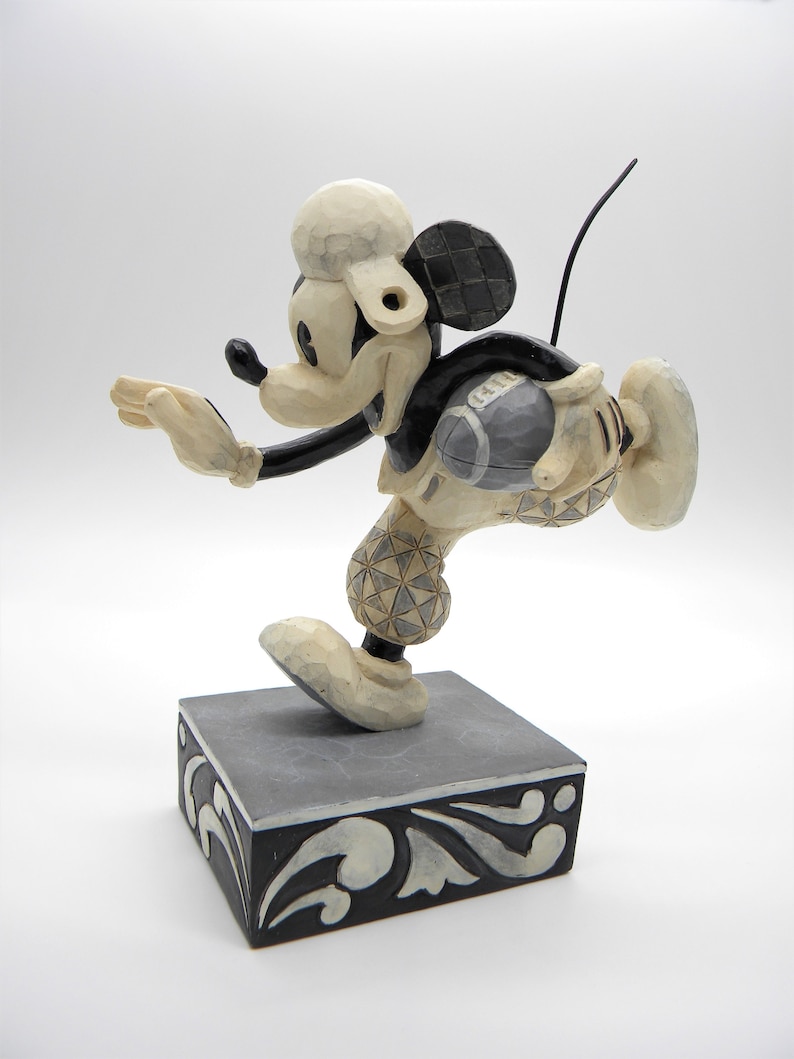 Puede incluir: Una figura tallada de Mickey Mouse en una pose din&aacute;mica, con una combinaci&oacute;n de colores crema y negro. La figura est&aacute; montada sobre una base cuadrada con adornos decorativos. El dise&ntilde;o de la figura presenta patrones geom&eacute;tricos y una est&eacute;tica caprichosa.